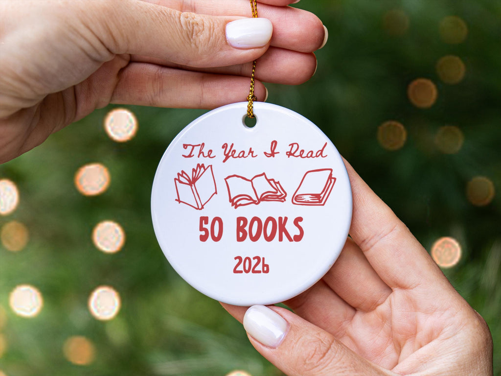 May include: A round white ceramic ornament featuring red text that reads The Year I Read 50 BOOKS 2026. The design includes three red line-art illustrations of books in various open and closed positions, suspended by a thin gold-coloured chain.