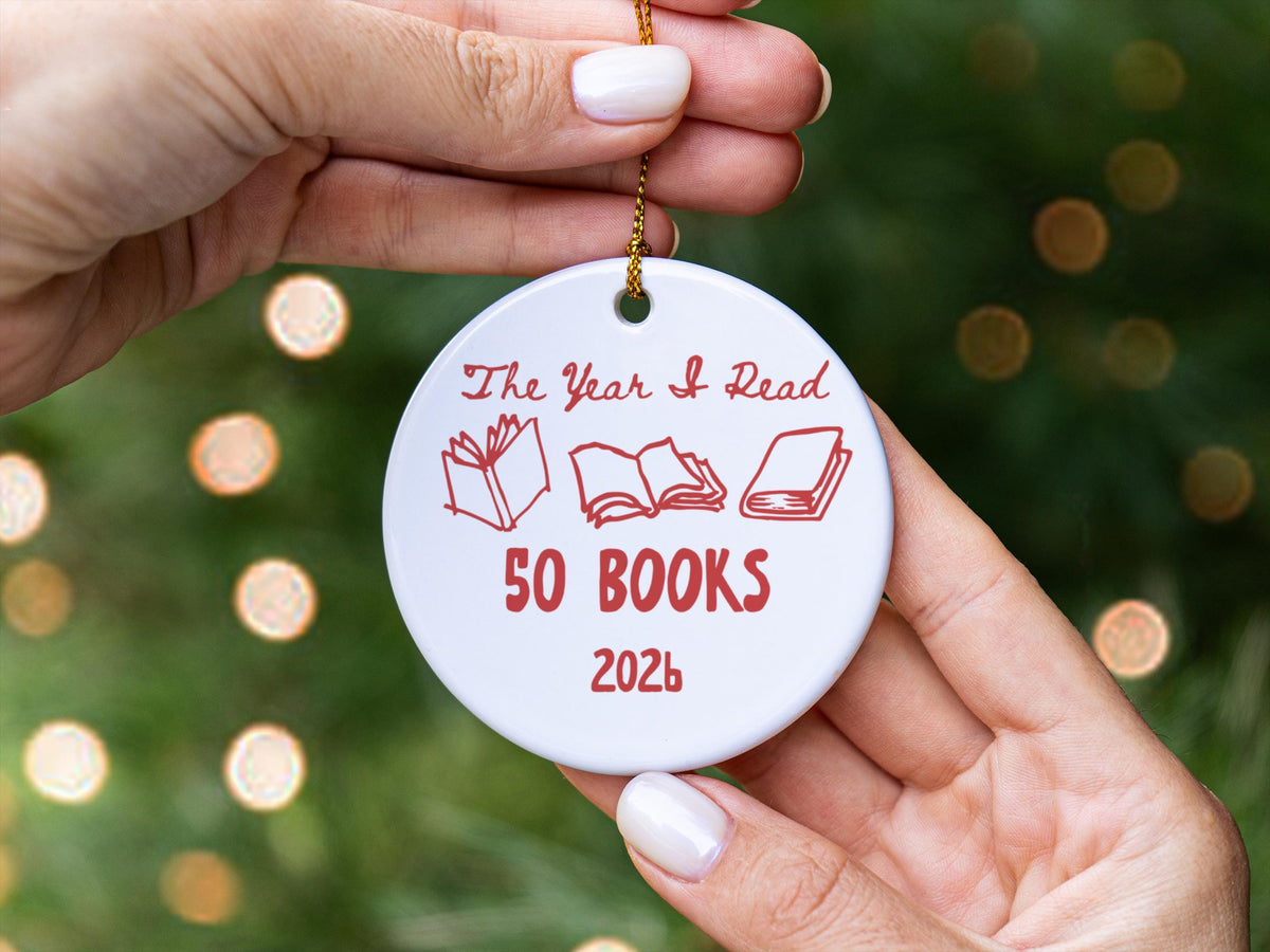 May include: A round white ceramic ornament featuring red text that reads The Year I Read 50 BOOKS 2026. The design includes three red line-art illustrations of books in various open and closed positions, suspended by a thin gold-coloured chain.