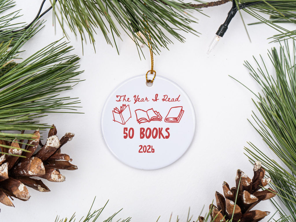 May include: A round white ceramic ornament featuring red text that reads The Year I Read 50 BOOKS 2026, accompanied by three red line-art illustrations of open and closed books, suspended by a thin gold string for hanging on a tree.