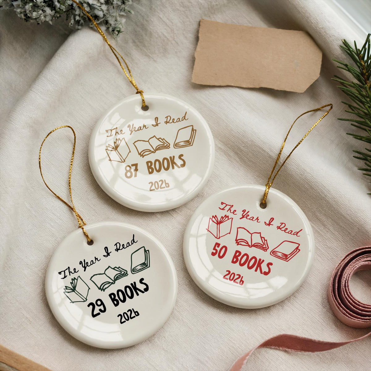 May include: Three round white ceramic ornaments with gold hanging strings. Each features the text "The Year I Read" above illustrations of open books, followed by a specific number of books and the year "2026" printed in gold, red, or black ink.