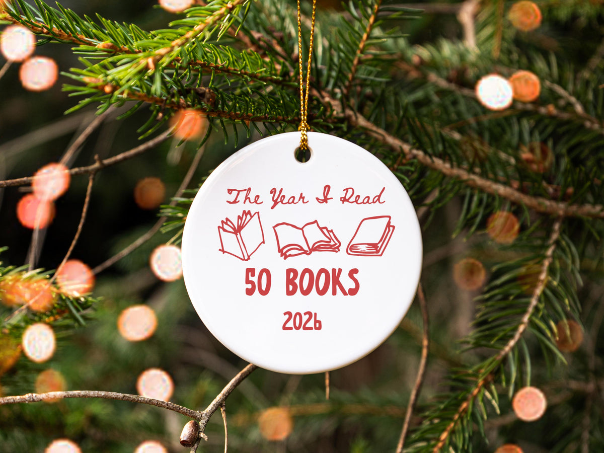 May include: A round white ceramic ornament hanging by a gold string. The front features red text that reads The Year I Read 50 BOOKS 2026, accompanied by three red line-art illustrations of open and closed books arranged in a horizontal row.